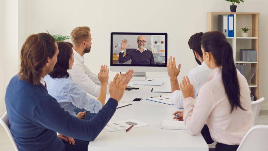 图片展示了几位职场人士通过视频会议与一位远程同事交流，他们正友好地挥手致意。会议环境看起来专业而整洁。